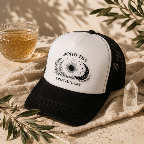 A black and white trucker hat with a 'Boho Tea Apothecary' graphic, styled on a beige fabric surface with a glass of tea, a crystal, and green leaves.