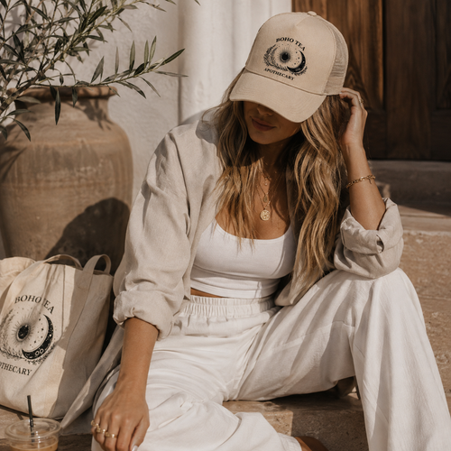 A woman sits outdoors wearing a beige trucker hat with a black 'BOHO TEA APOTHECARY' logo, a white crop top, a light beige button-up shirt, and white pants. A matching beige tote bag with the same logo sits beside her on stone steps.